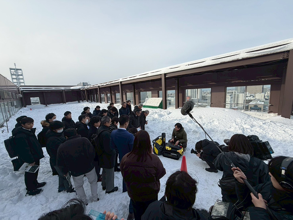 見学会の様子。自動除雪ロボットの周りに人や報道カメラなどが集まっている。