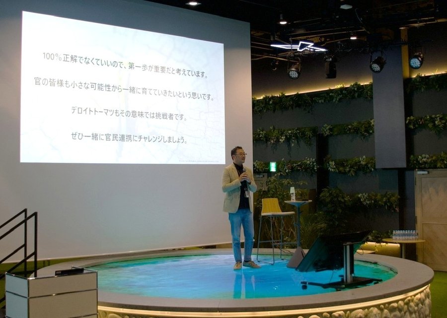 開会挨拶で、スライドを背景にマイクを持って話す伊東真史氏。